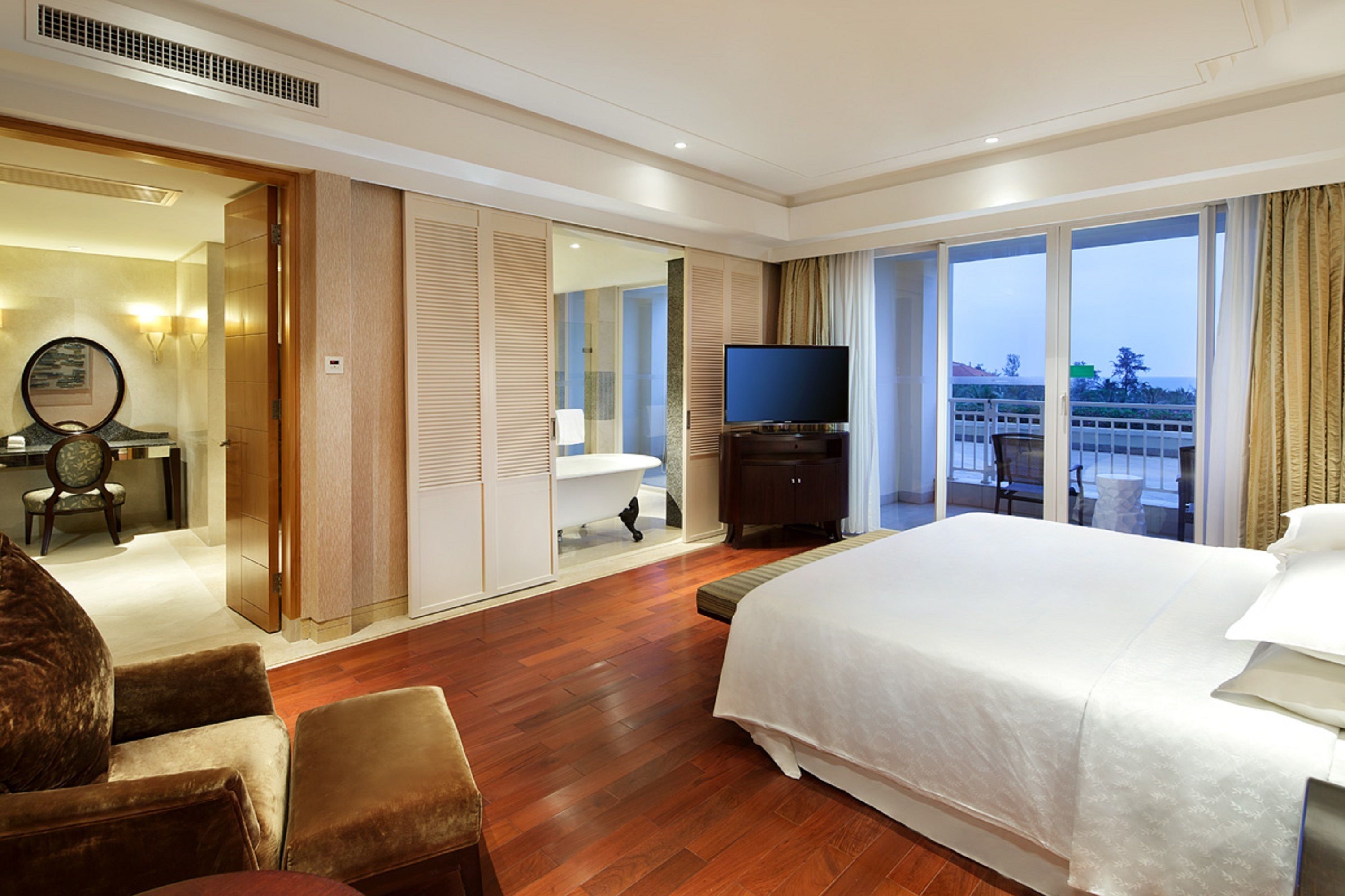 Habitación de hotel con una cama grande, televisión, baño con bañera y vistas a la ciudad a través de amplios ventanales.