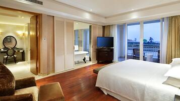 Habitación de hotel con una cama grande, televisión, baño con bañera y vistas a la ciudad a través de amplios ventanales.