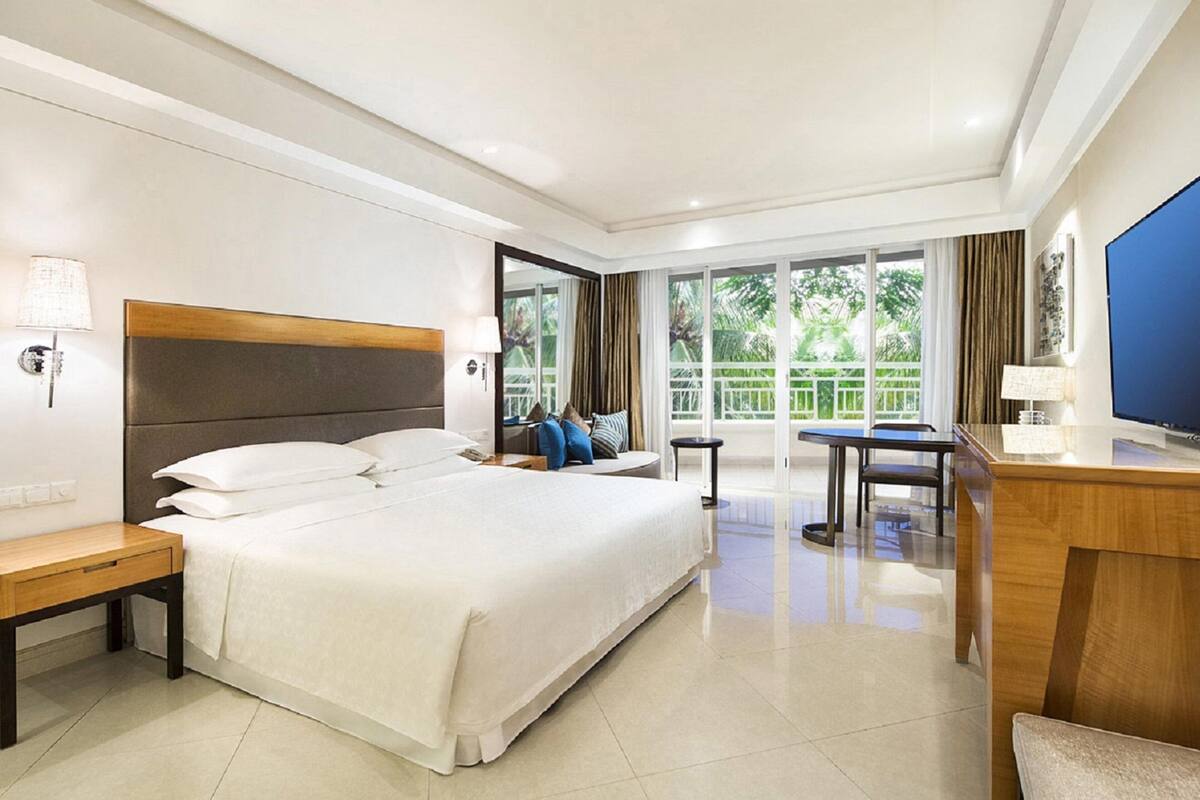 Habitación de hotel con una cama grande, un escritorio, una silla, un televisor y un balcón con vistas a la vegetación.