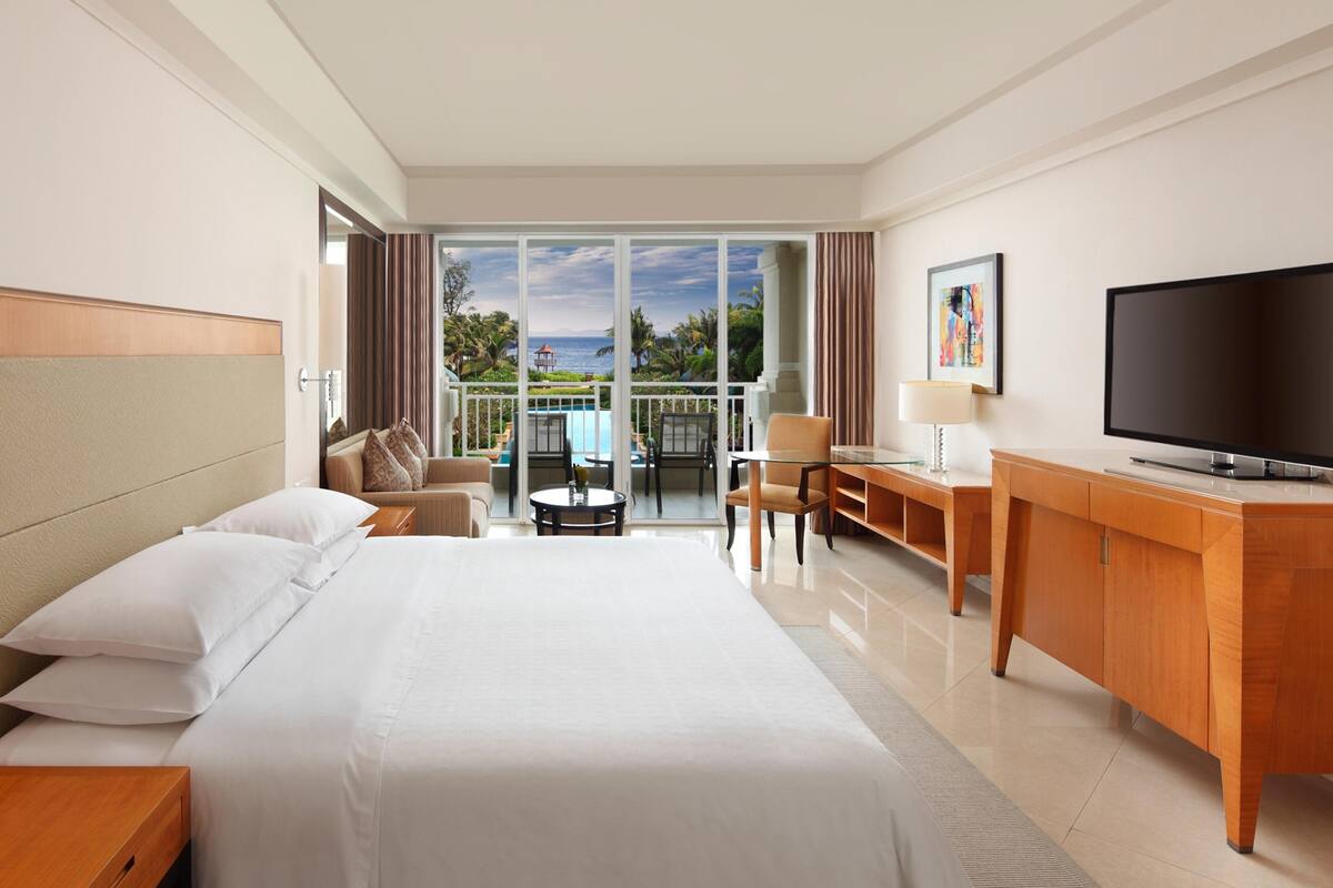 Habitación de hotel con cama, escritorio, televisor y balcón con vista a la piscina.