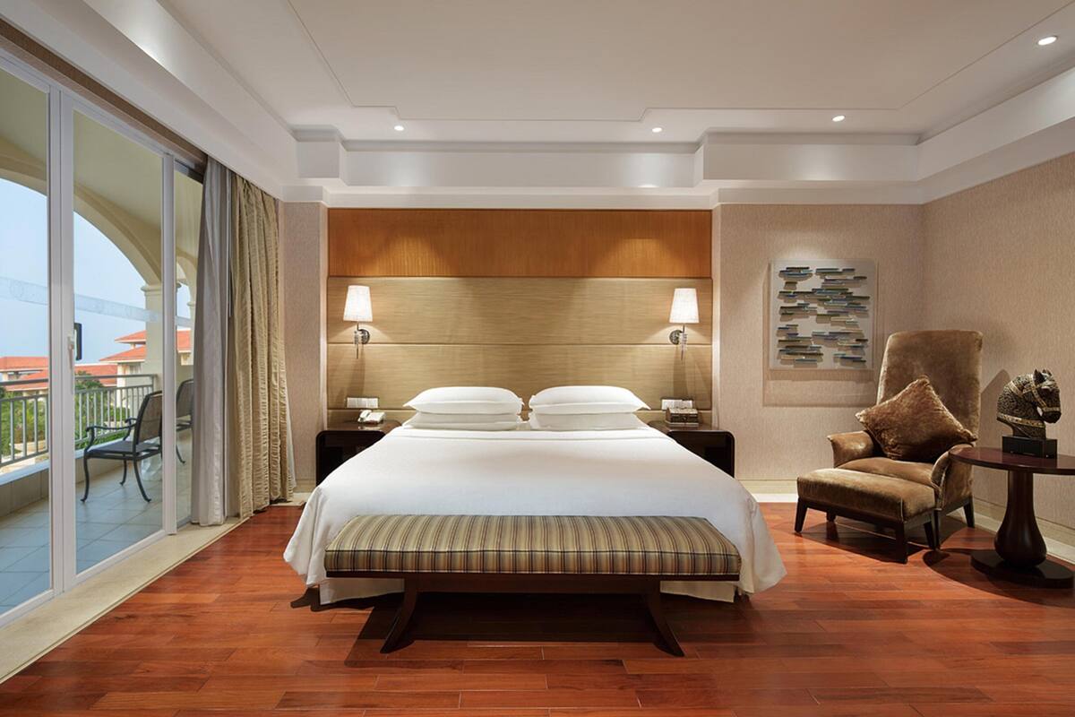 Habitación de hotel con una cama grande, dos mesitas de noche, una silla, una mesita pequeña y un balcón con una silla.