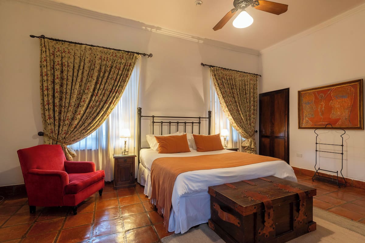 A bedroom with a bed, a red armchair, a wooden chest, and curtains.
