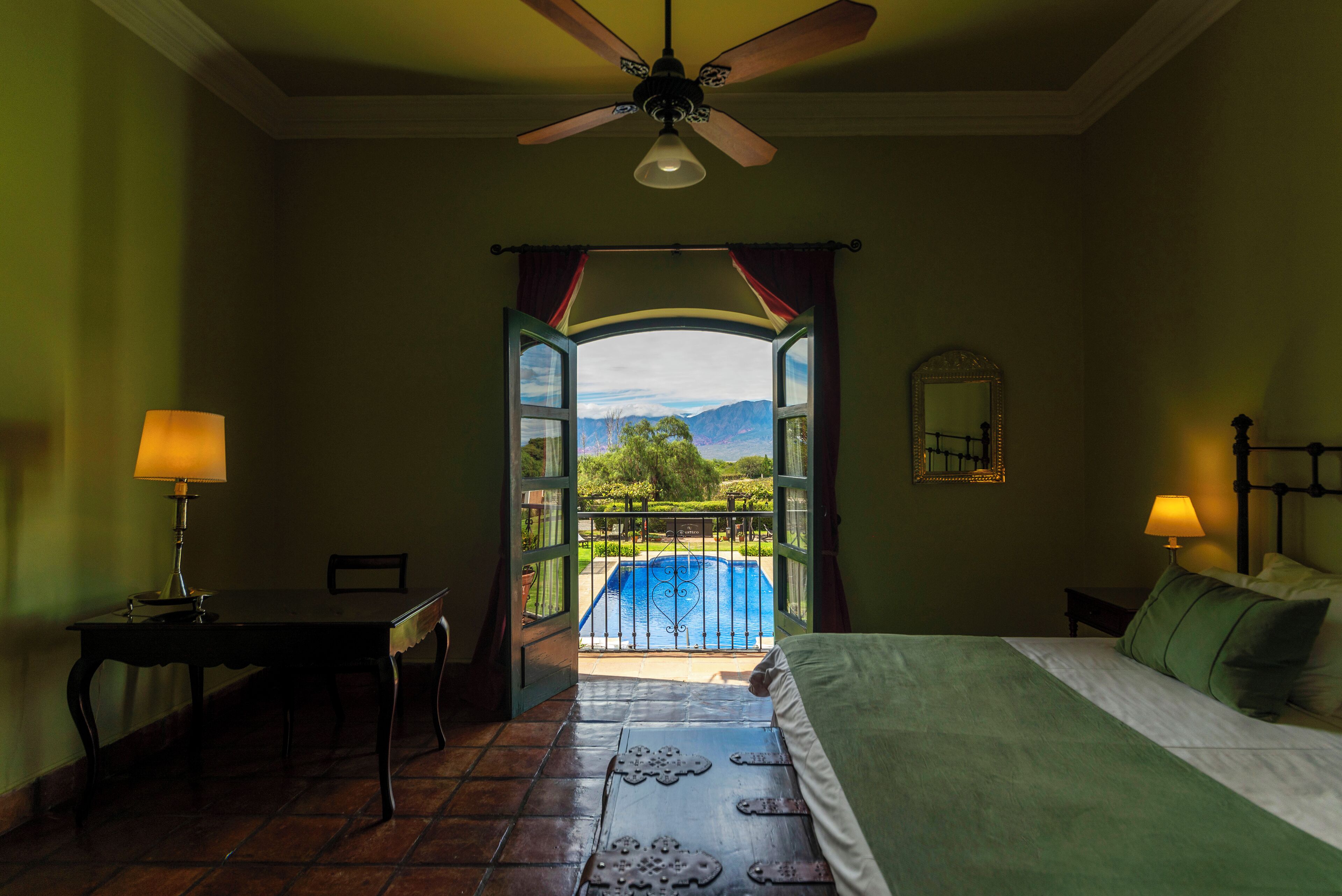 Ein Schlafzimmer mit Bett, Schreibtisch, Stuhl, Deckenventilator und Blick auf einen Swimmingpool und Berge durch eine offene Tür.