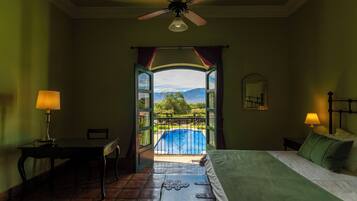 Ein Schlafzimmer mit Bett, Schreibtisch, Stuhl, Deckenventilator und Blick auf einen Swimmingpool und Berge durch eine offene Tür.