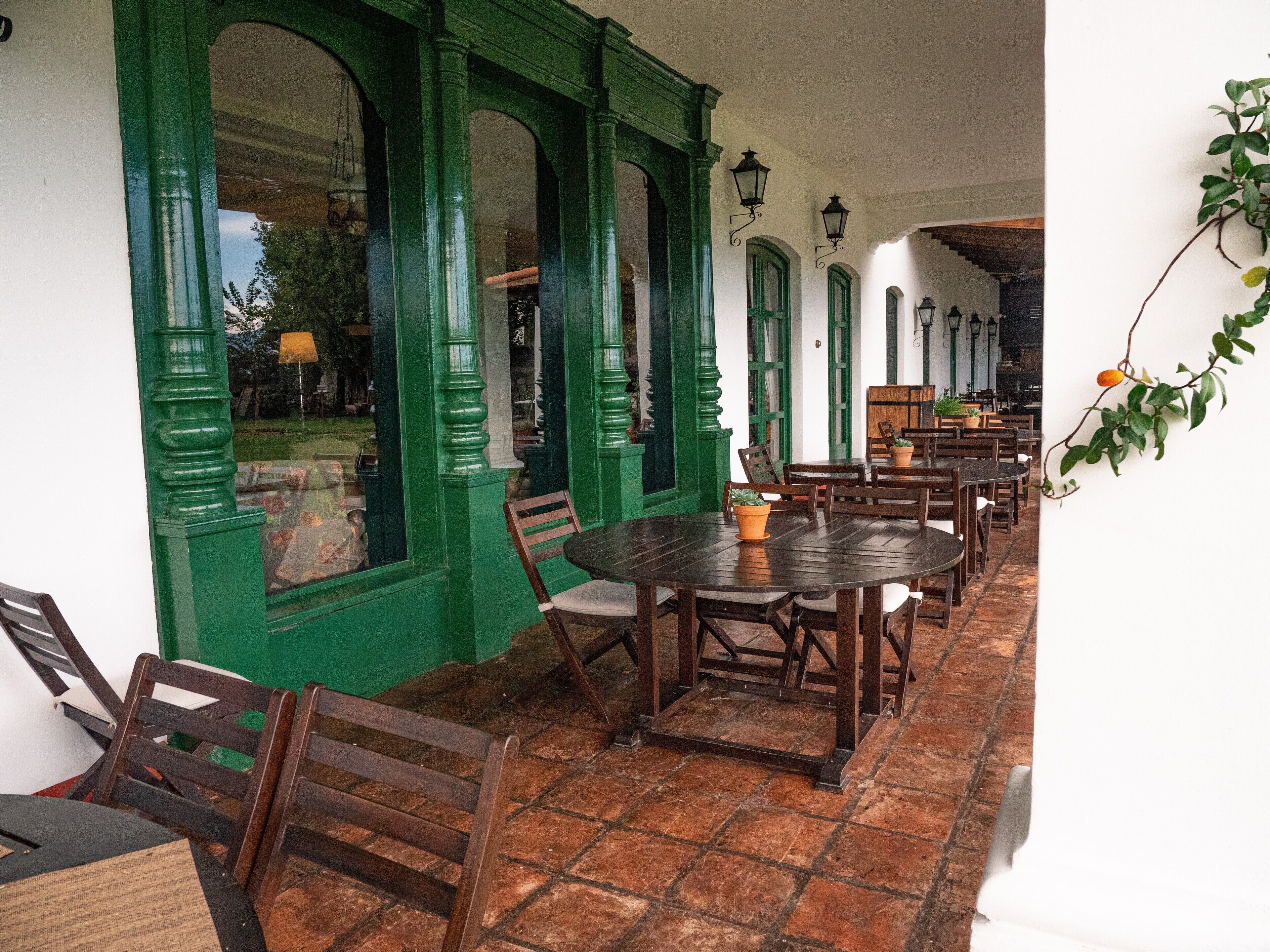 Un patio cubierto con ventanas de marco verde, mesas y sillas de madera y suelo de baldosas.