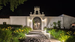 Un edificio blanco con una entrada central en arco y una torre de campanas, rodeado de jardines cuidados y un camino de grava.