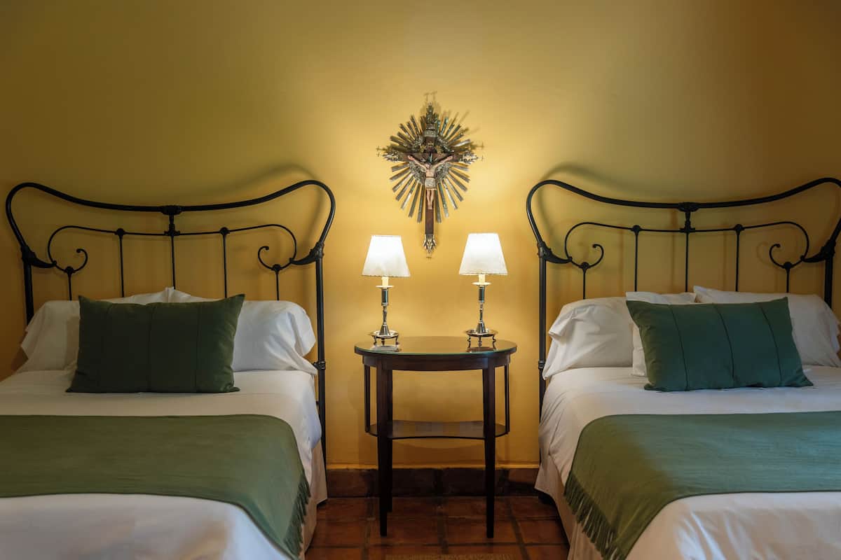 A hotel room with two beds, each with a green bedspread and white pillows. There is a small table with two lamps between the beds.