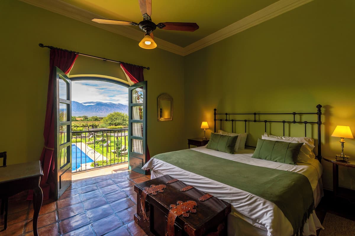 A bedroom with a bed, a nightstand, a lamp, a wooden chest, and a view of a pool and mountains through an open door.