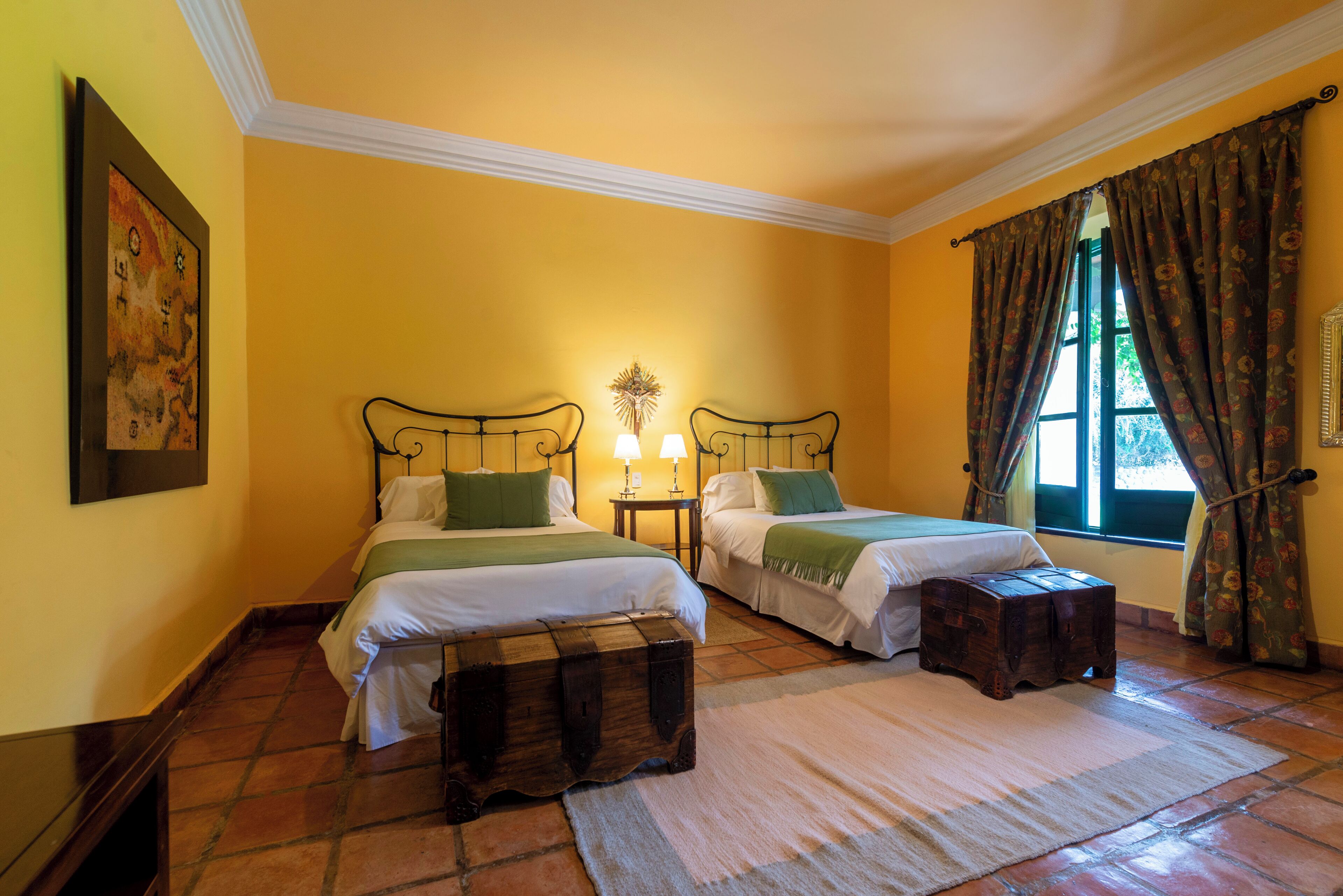 A room with two beds, a wooden chest, a rug, and a painting on the wall.