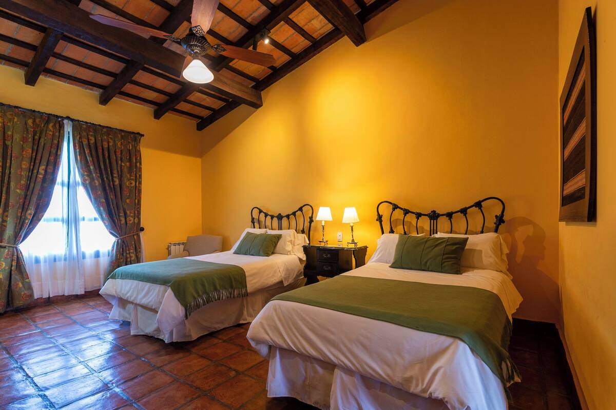 A room with two beds, wooden beams on the ceiling, and a window with curtains.