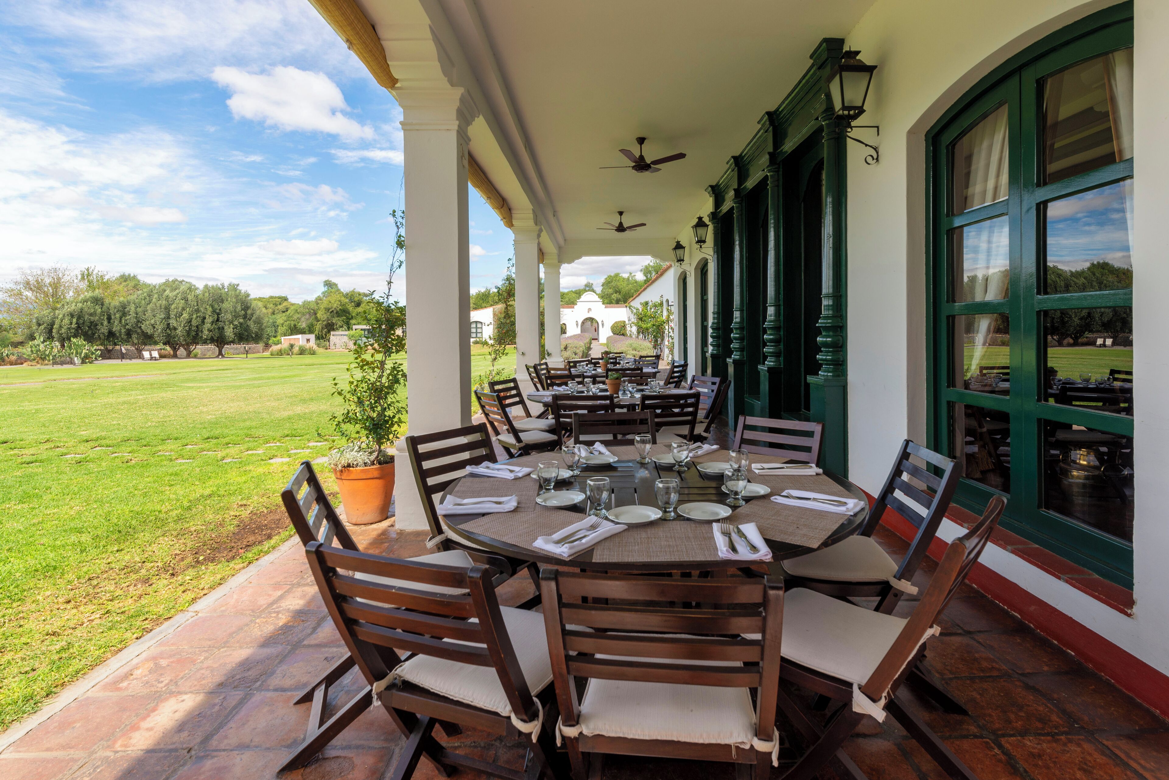 Un área de comedor al aire libre techada con mesas y sillas redondas, vista a un césped verde y un edificio al fondo.