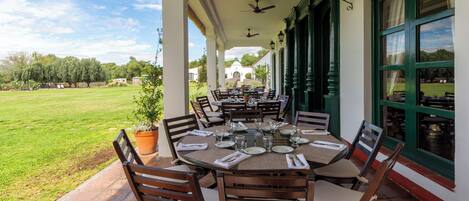 Un área de comedor al aire libre techada con mesas y sillas redondas, vista a un césped verde y un edificio al fondo.