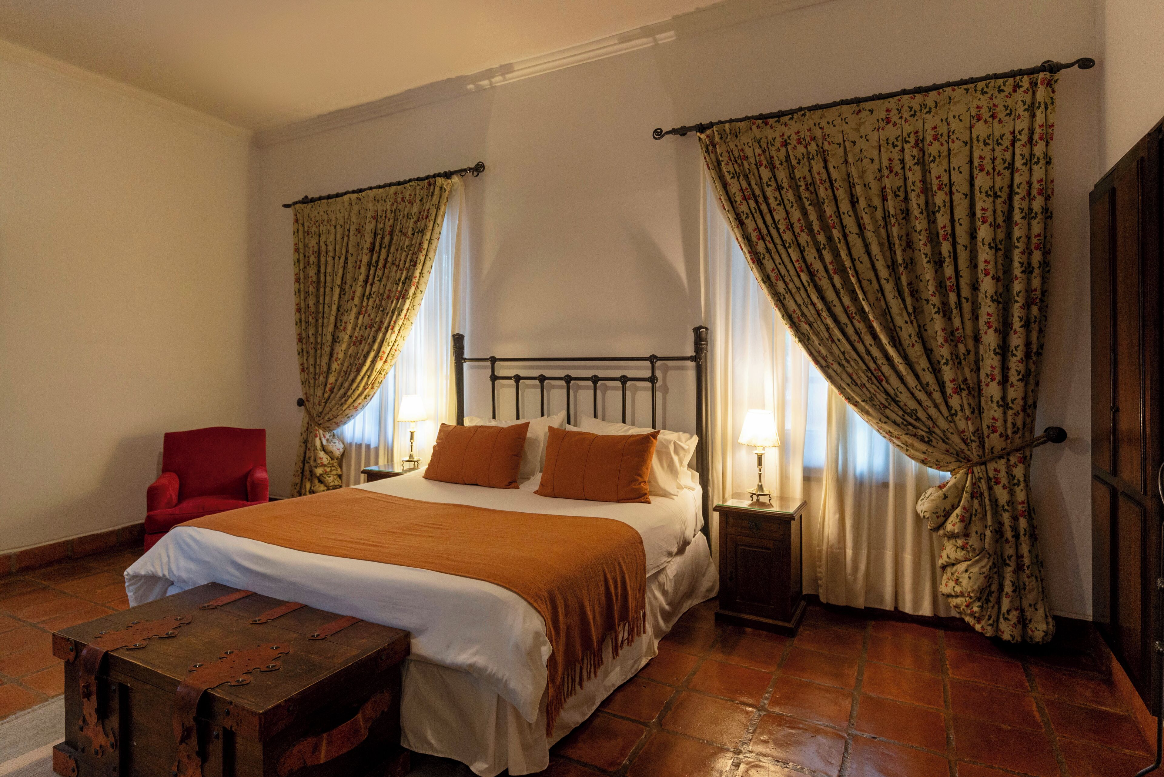 Un dormitorio con una cama grande, dos mesitas de noche, un banco de madera, una silla roja y cortinas florales.