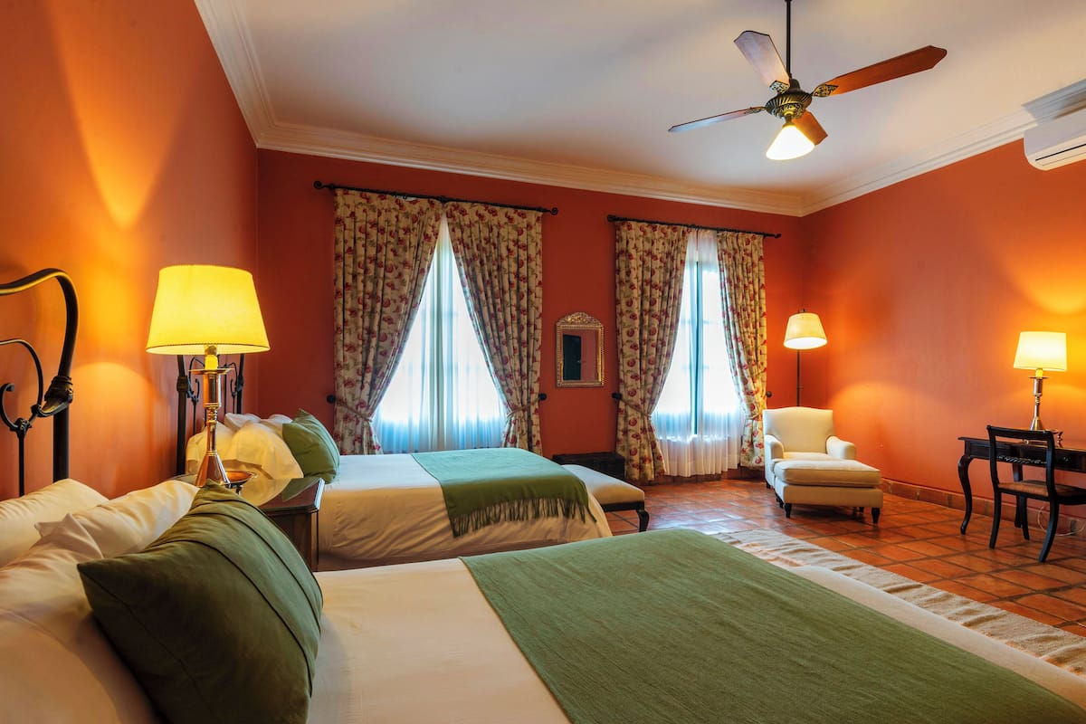 A hotel room with two beds, a chair, a desk, and a ceiling fan.