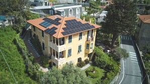 A yellow building with a red-tiled roof and solar panels. It has a balcony and is surrounded by greenery.