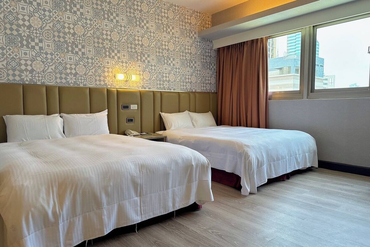A hotel room with two beds, a patterned wall, a headboard with lights, a window with curtains, and a city view.