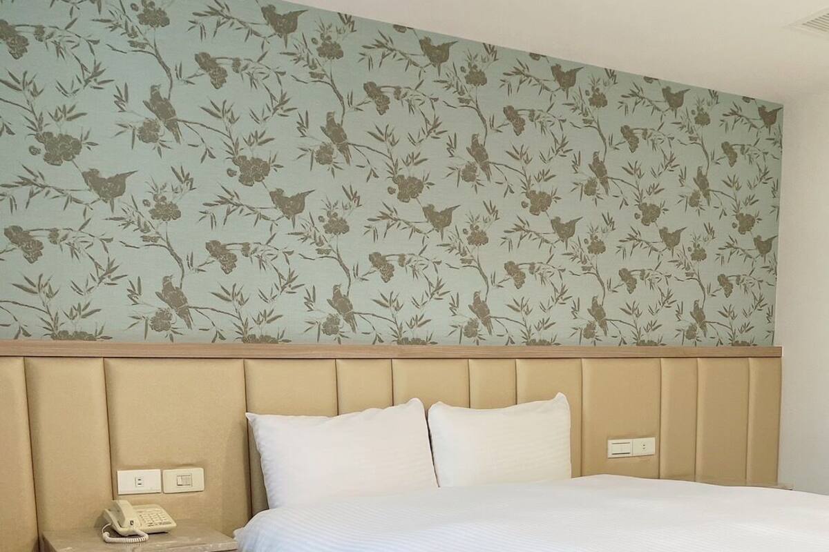 A hotel room with a bed, a nightstand with a phone, and a floral wallpaper.