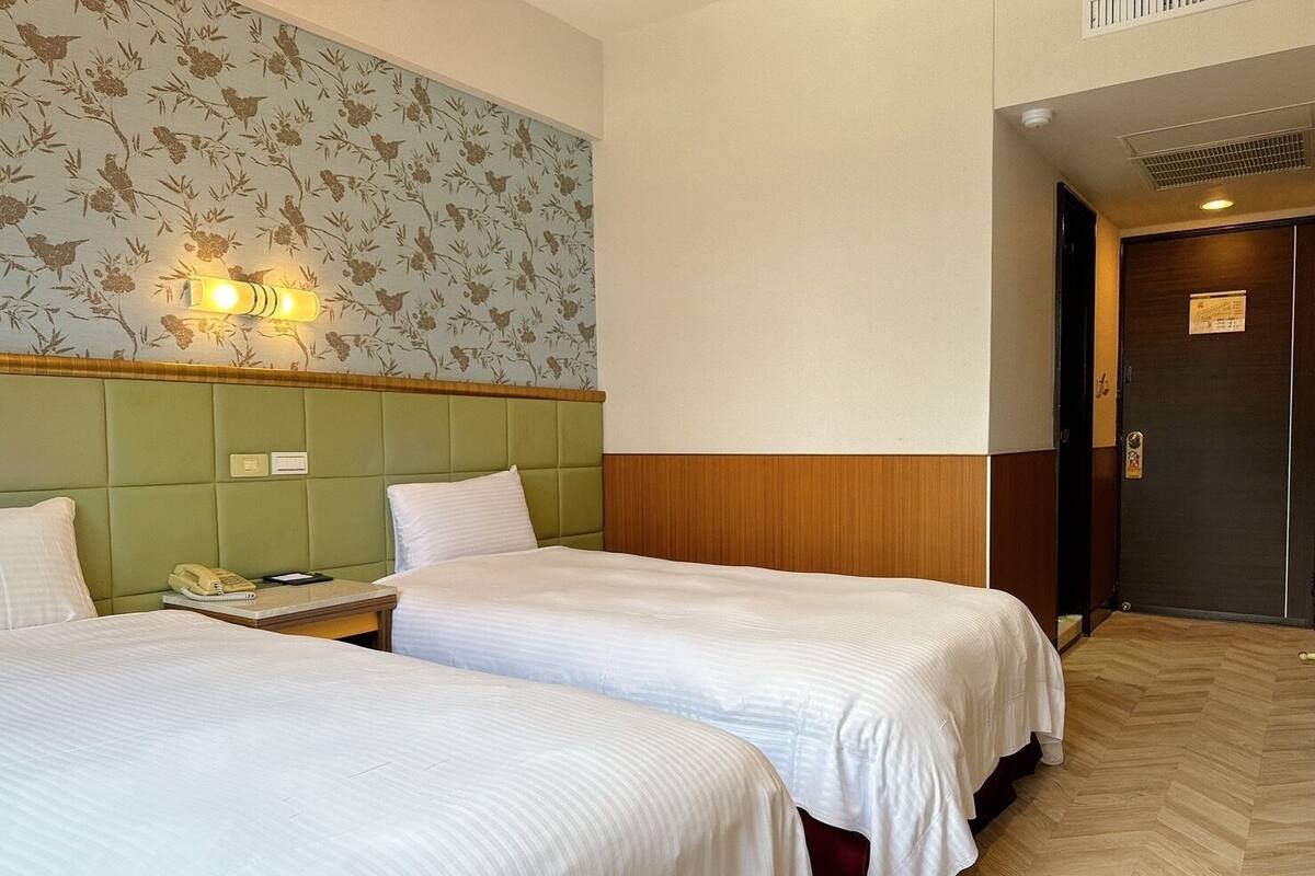 A hotel room with two beds, a green headboard, a floral wallpaper, and a wooden door.