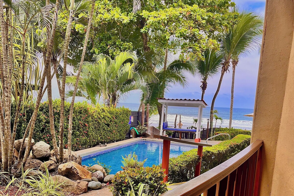A pool area with a view of the ocean, surrounded by tropical plants and a wooden deck.