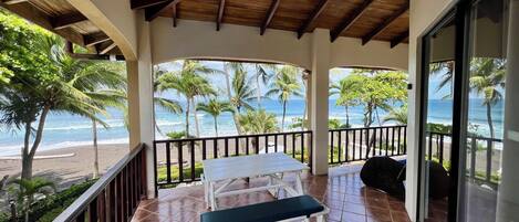 A covered outdoor area with a table and benches, overlooking a beach with palm trees and the ocean.