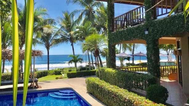 A poolside area with a view of the beach, palm trees, and a covered seating area. - The Backyard Hotel (Jacó)