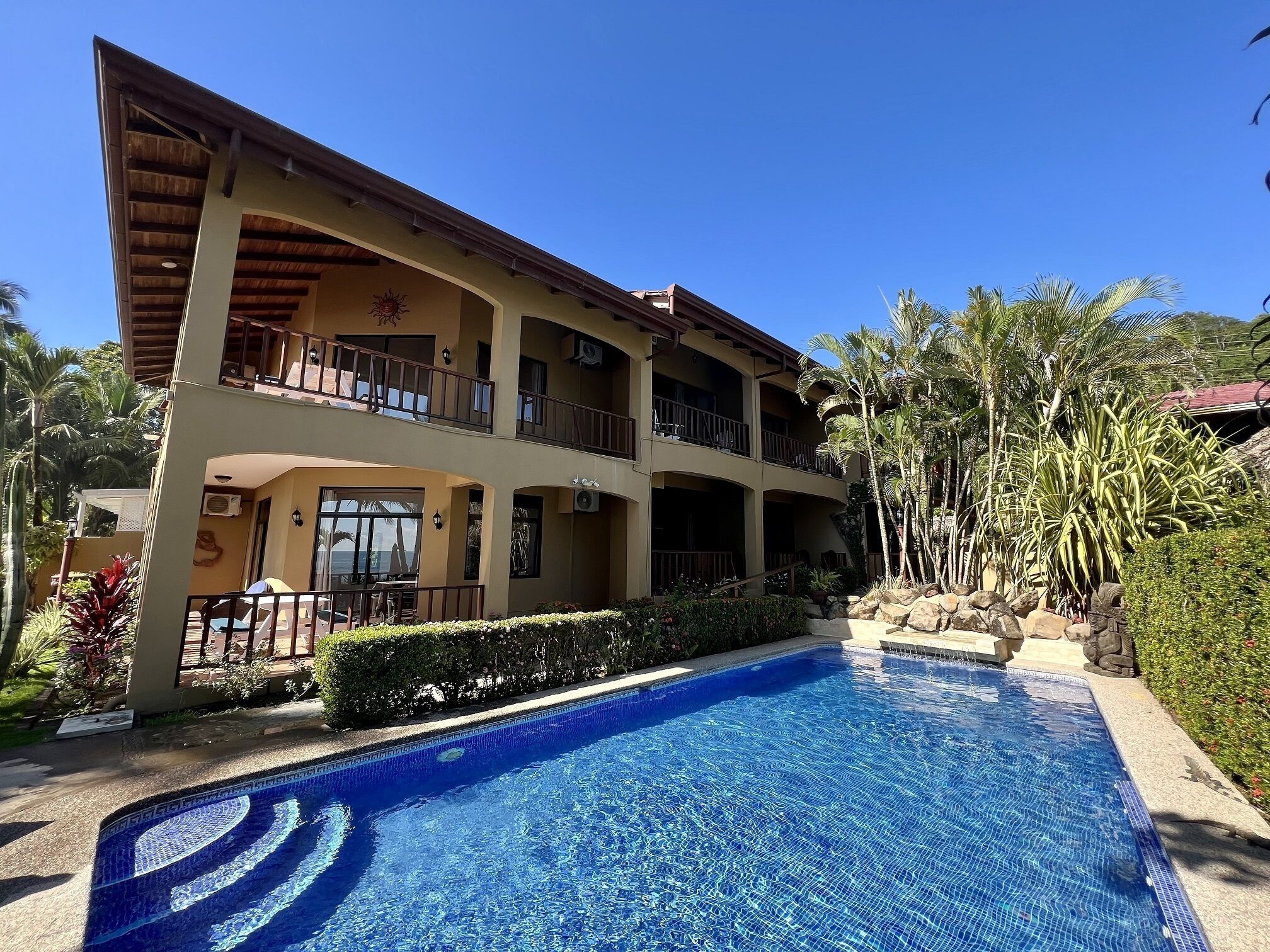 A two-story hotel with a swimming pool, balconies, and tropical landscaping.