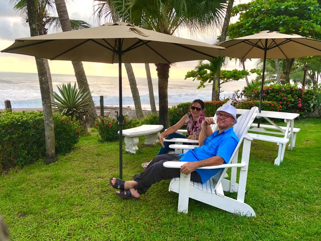 Two people relaxing on white outdoor chairs with beige umbrellas, overlooking a beach with palm trees and a view of the ocean.