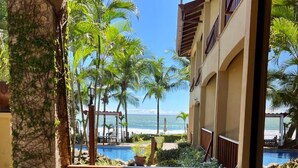 A balcony with a view of a pool and beach, wooden furniture, and a vase with flowers. - The Backyard Hotel (Jacó)