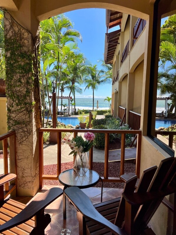 A balcony with a view of a pool and beach, wooden furniture, and a vase with flowers. - The Backyard Hotel (Jacó)