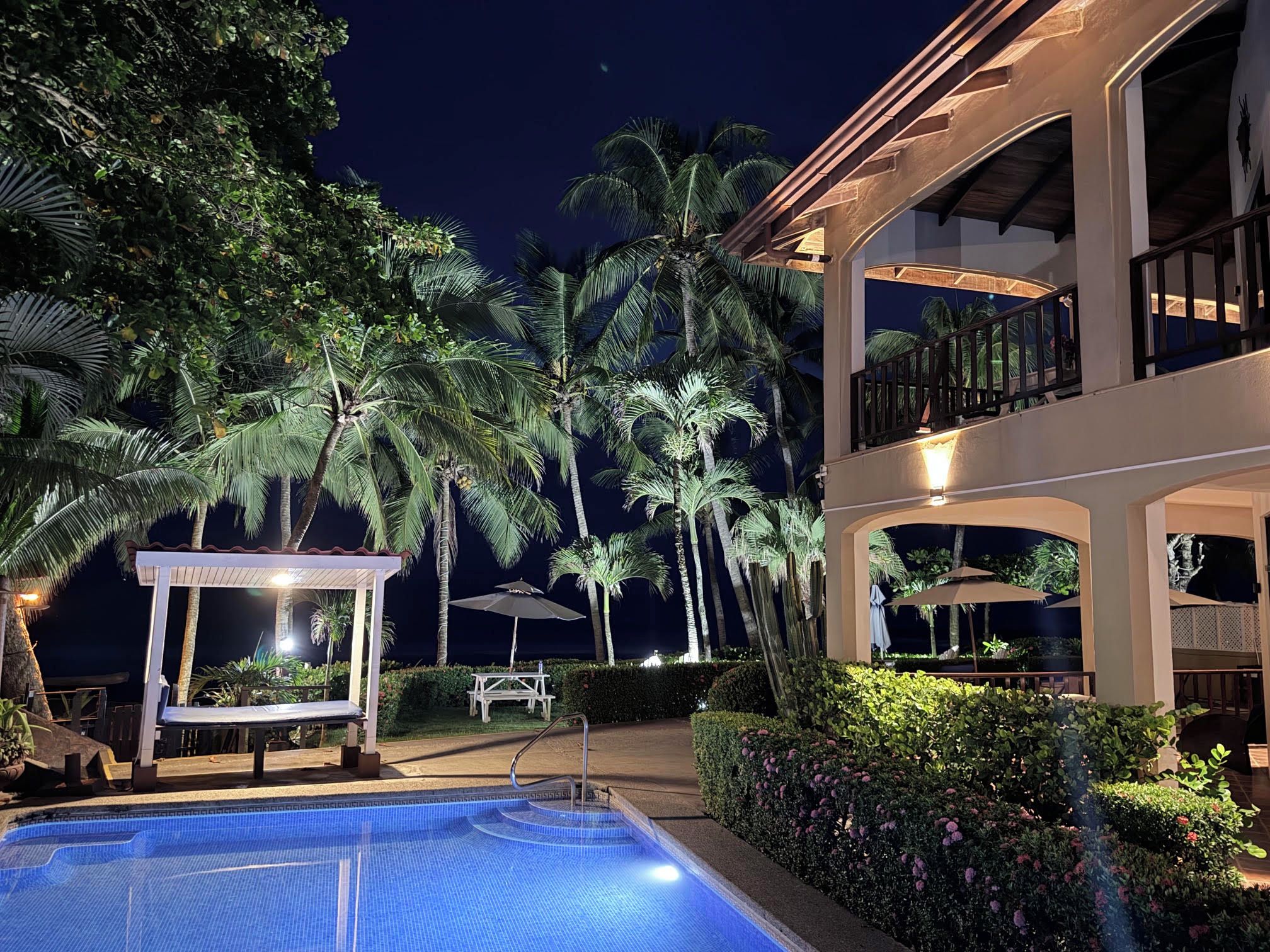 Área de piscina à noite, com espaço de estar coberto e um edifício com varandas.