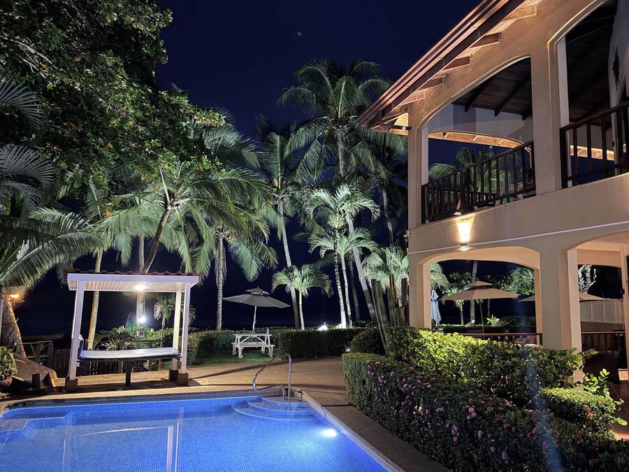 Área de piscina à noite, com espaço de estar coberto e um edifício com varandas.