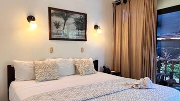 A hotel room with a large bed, a ceiling fan, an air conditioning unit, a painting on the wall, and a view of the outdoors through a window with curtains.
