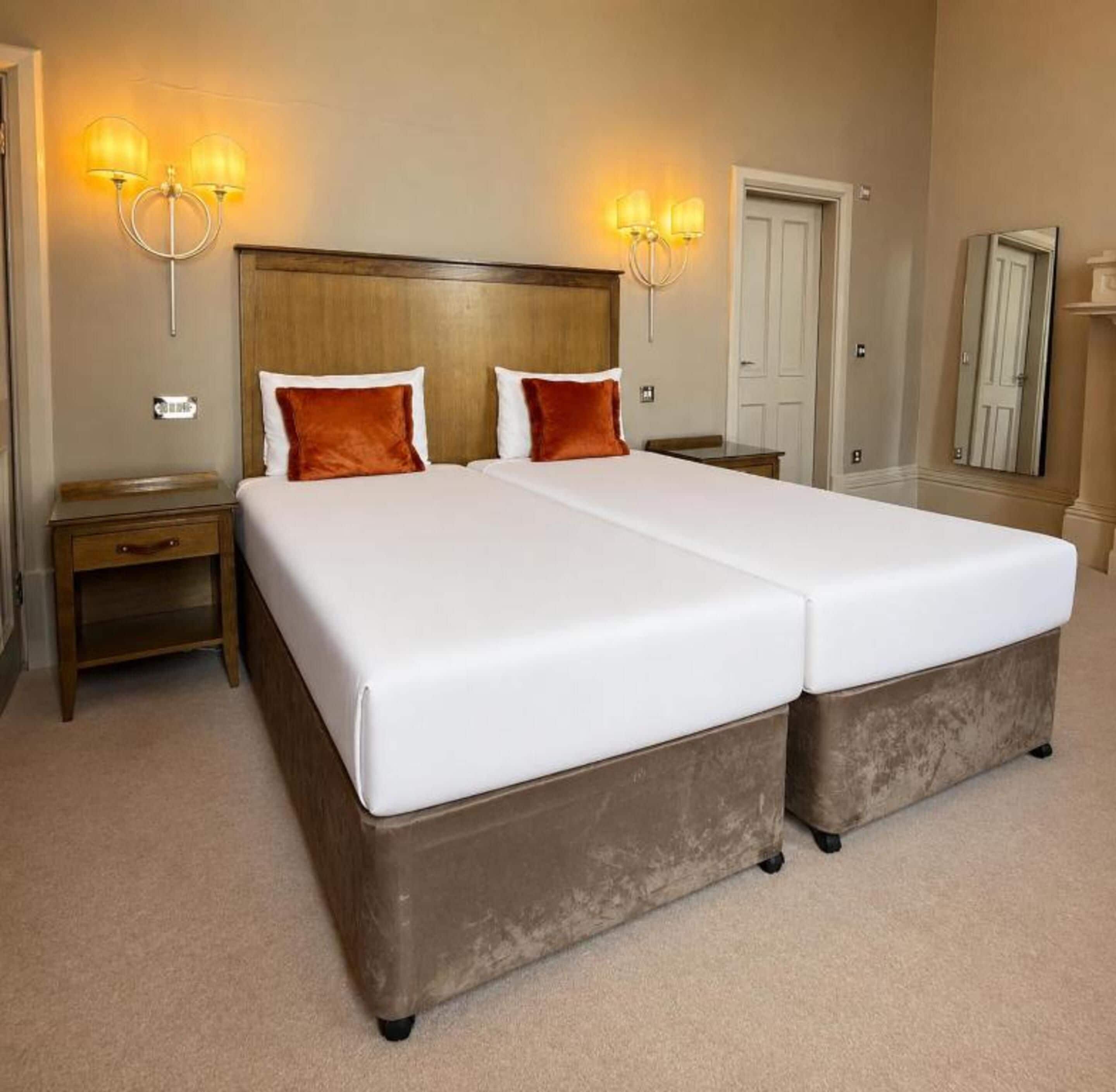 A double bed with two pillows, a nightstand, a wall-mounted lamp, and a door in a hotel room.