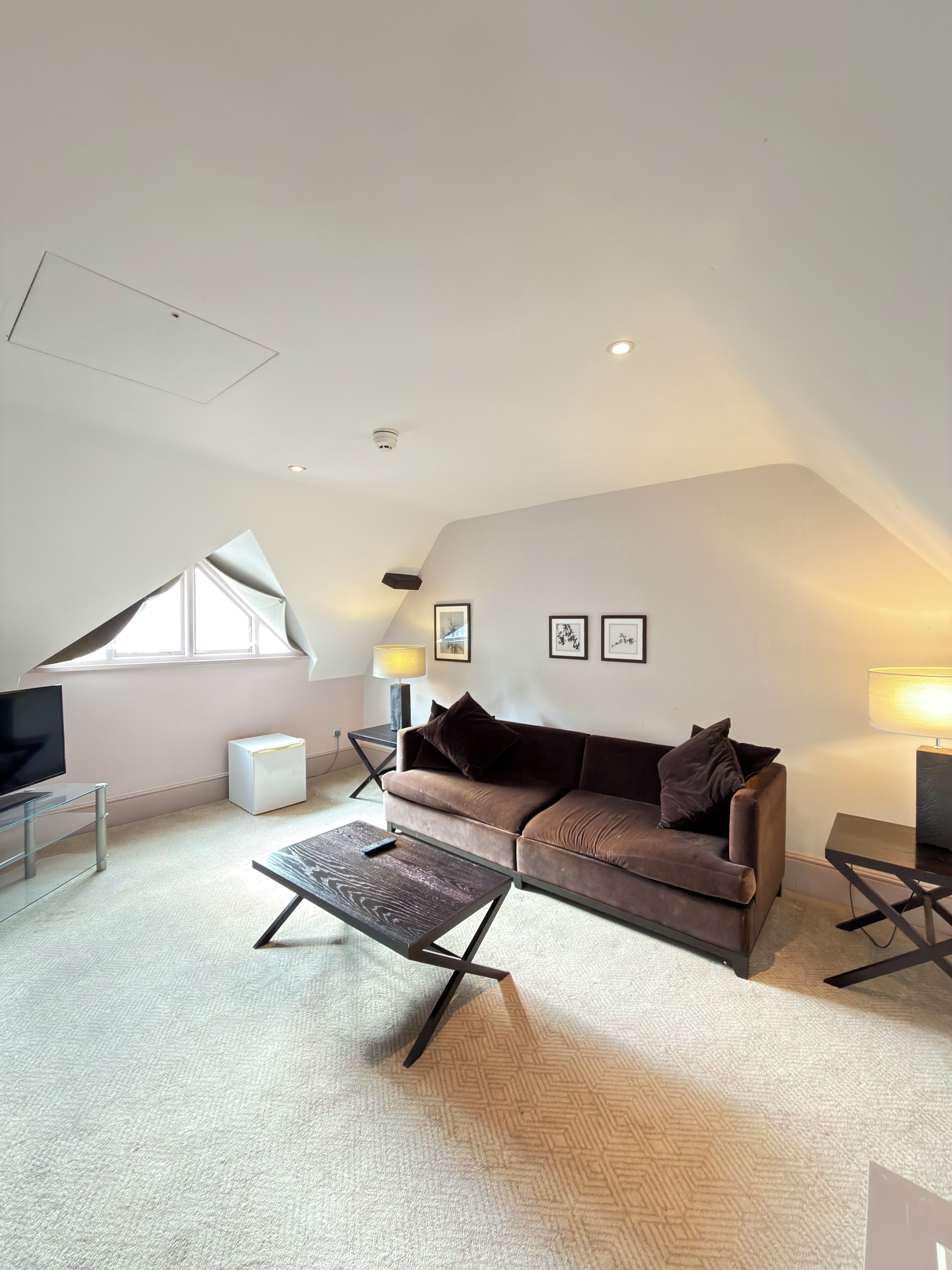 A modern living room with a brown sofa, a glass coffee table, a flat-screen TV, and a white mini-fridge.
