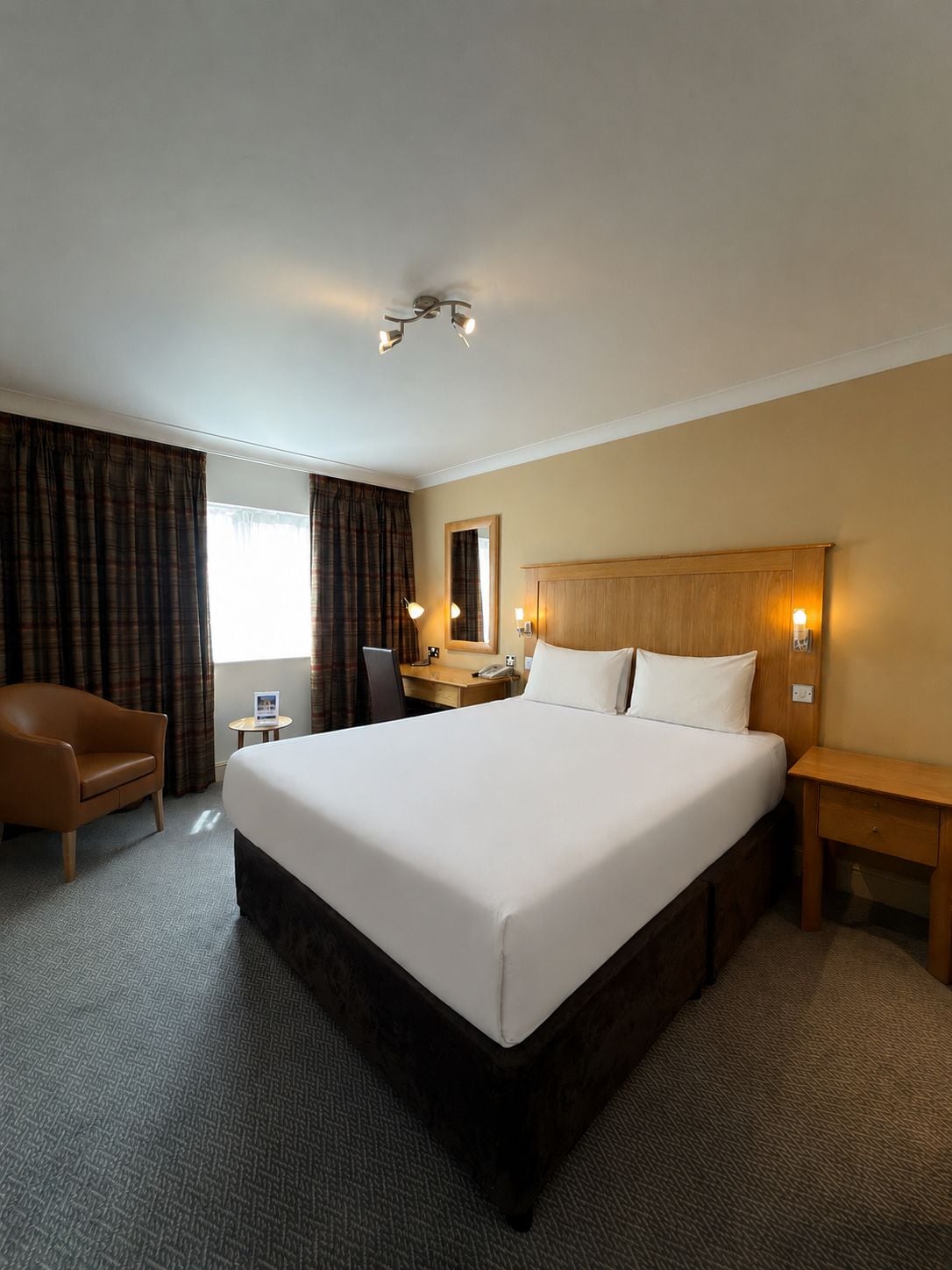 A hotel room with a large bed, a desk, a chair, a bedside table, and a mirror.