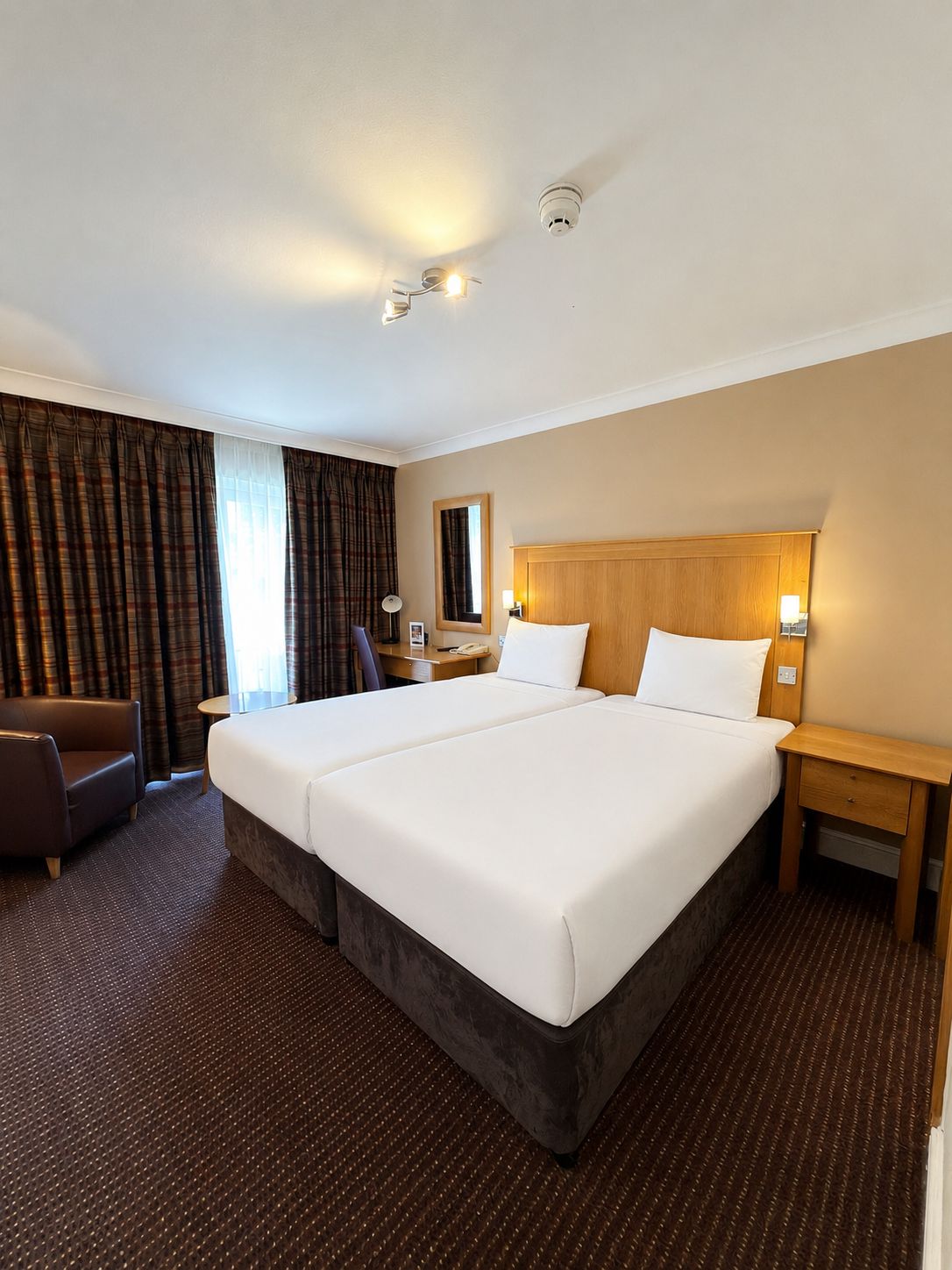 A hotel room with a large bed, a desk, a chair, a nightstand, a mirror, and a window with curtains.