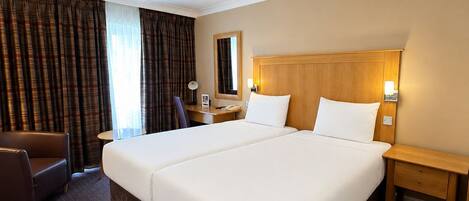 A hotel room with a large bed, a desk, a chair, a nightstand, a mirror, and a window with curtains.