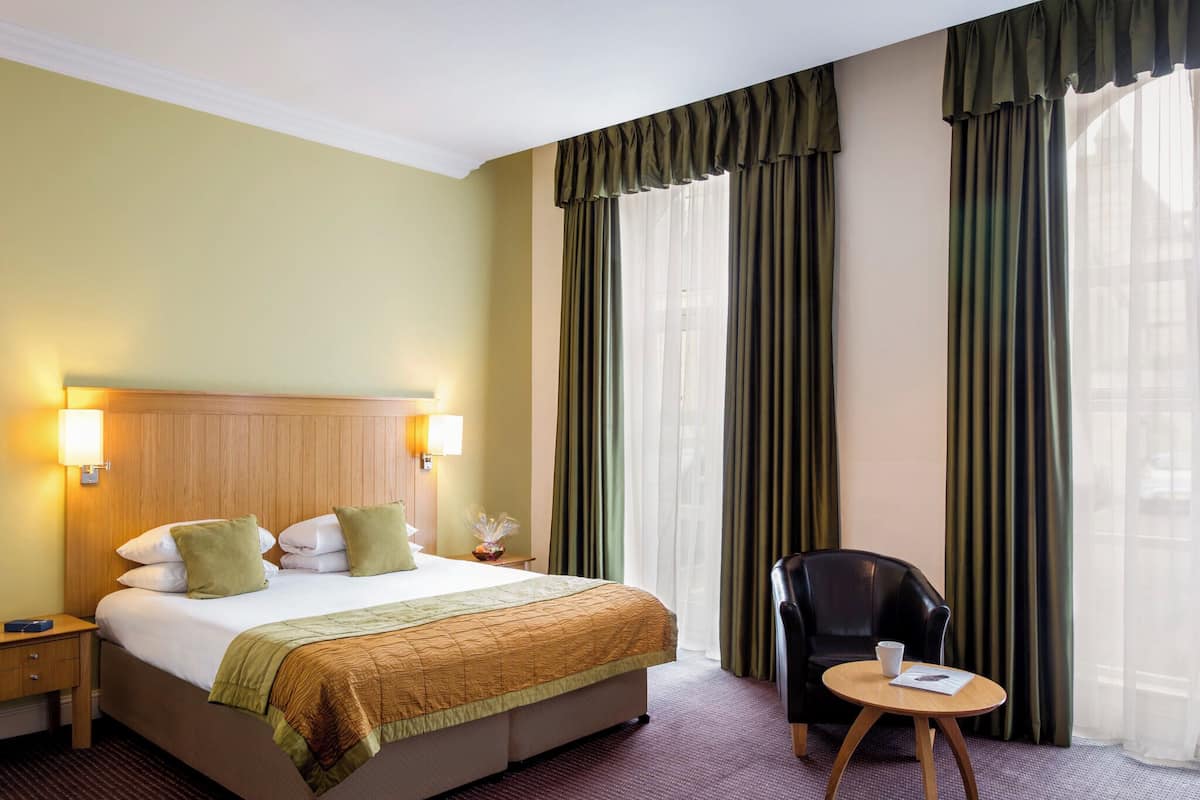 Hotellihuone, jossa on suuri sänky, kaksi yöpöytävalaisinta, tuoli, pieni pöytä ja näkymä kaupunkiin ohuiden verhojen läpi.