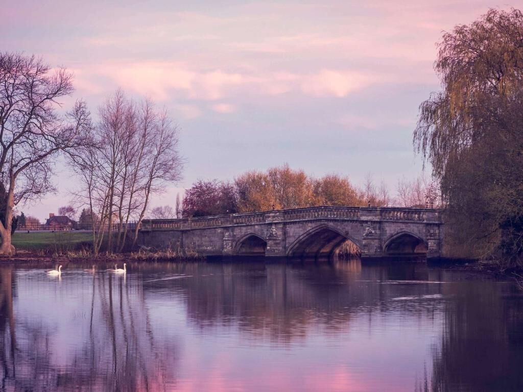 Un pont de pierre enjambe une rivière paisible où nagent des cygnes, le tout étant entouré d’arbres et surplombé par un ciel dégagé.