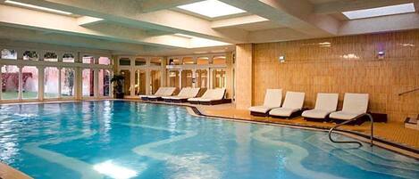 An indoor swimming pool with clear water, sunlit ceiling, and lounge chairs along the side.
