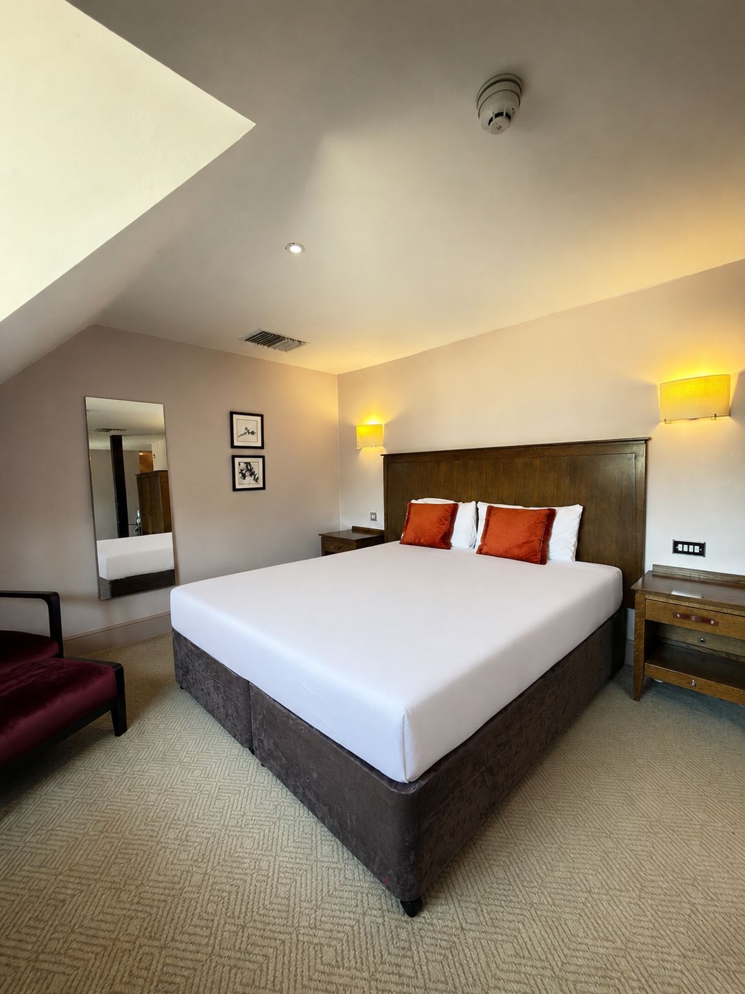A hotel room with a large bed, a nightstand, a bedside table, a chair, a mirror, and a wall-mounted light fixture.