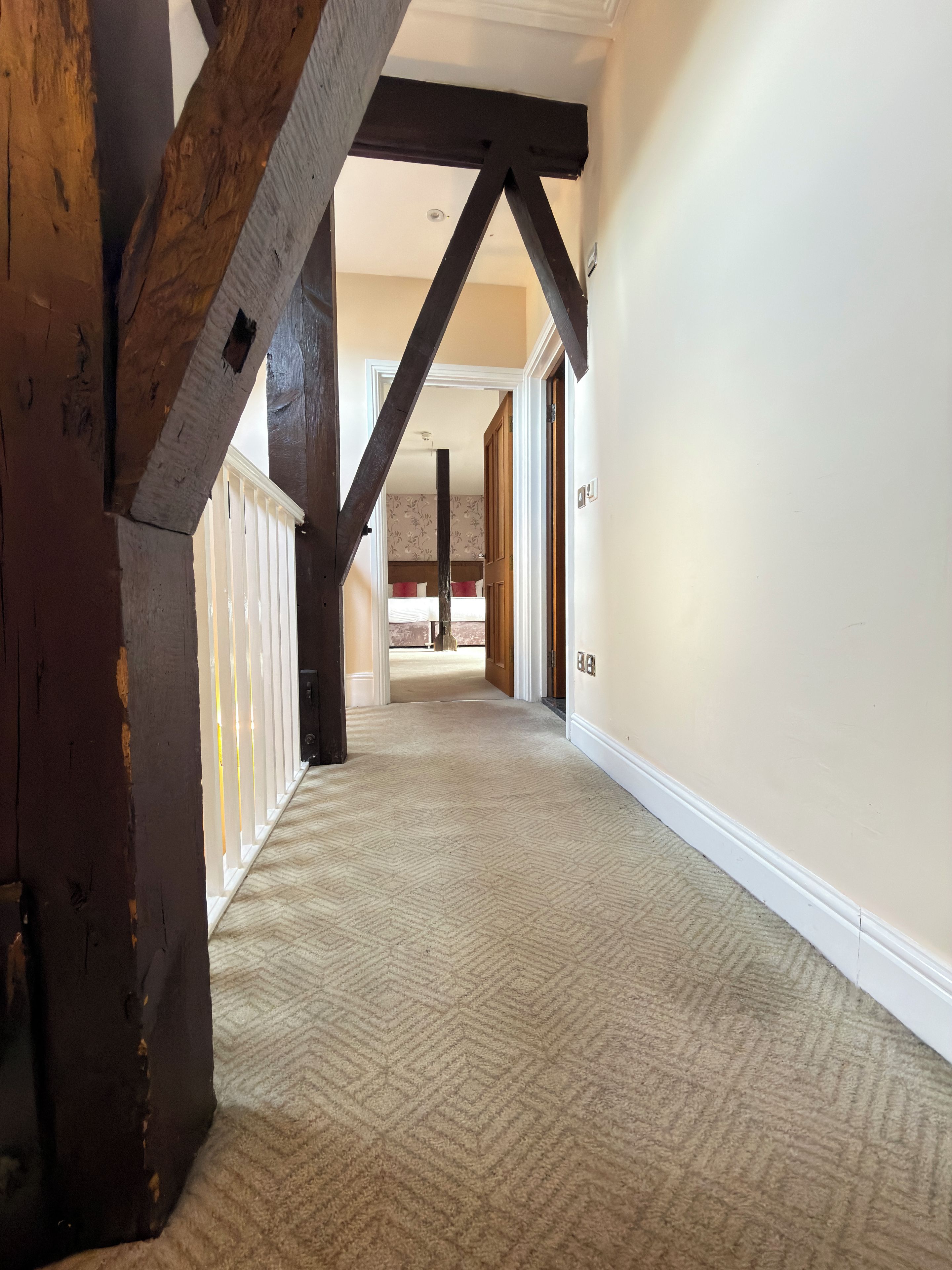 A hallway with a herringbone carpet, wooden beams, and a white door leading to a bedroom.