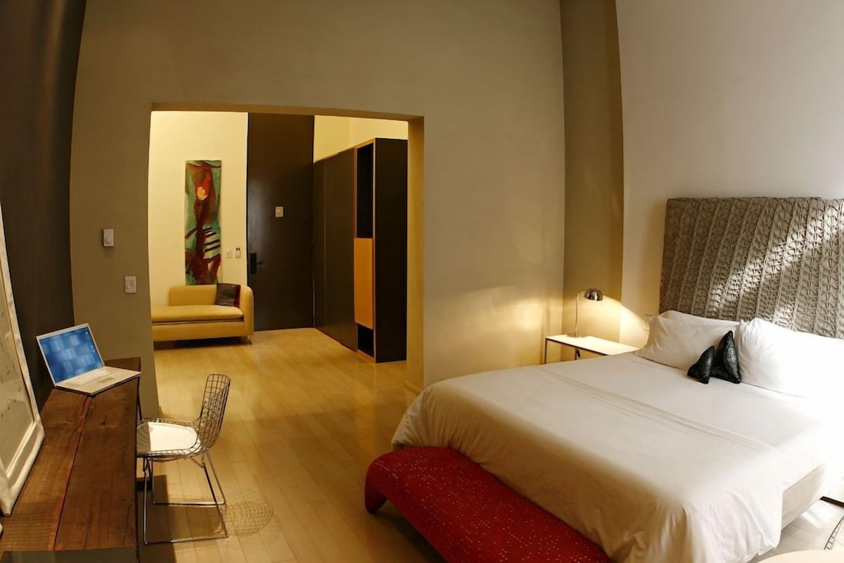 Une chambre d’hôtel avec un lit, un bureau avec un ordinateur portable, une chaise et un banc rouge.