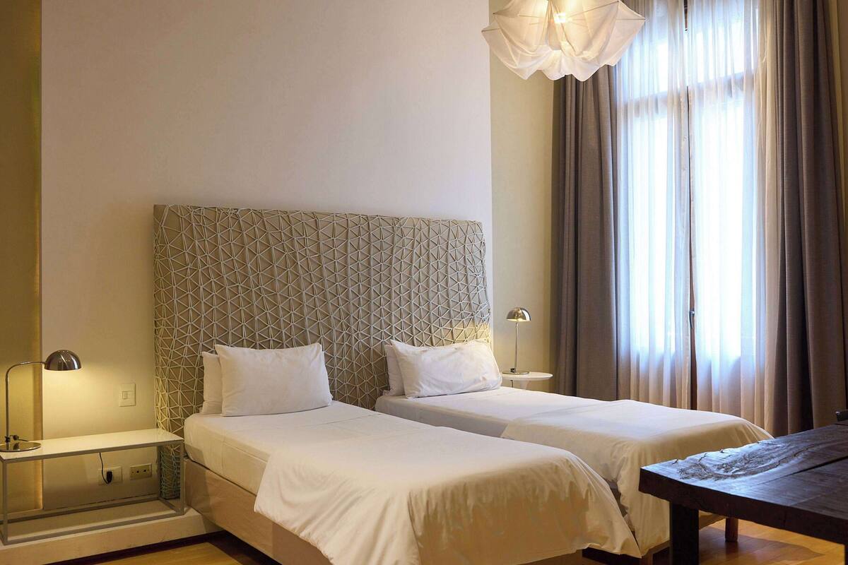 Une chambre d’hôtel avec deux lits, une table en bois, une lampe et une fenêtre avec des rideaux.