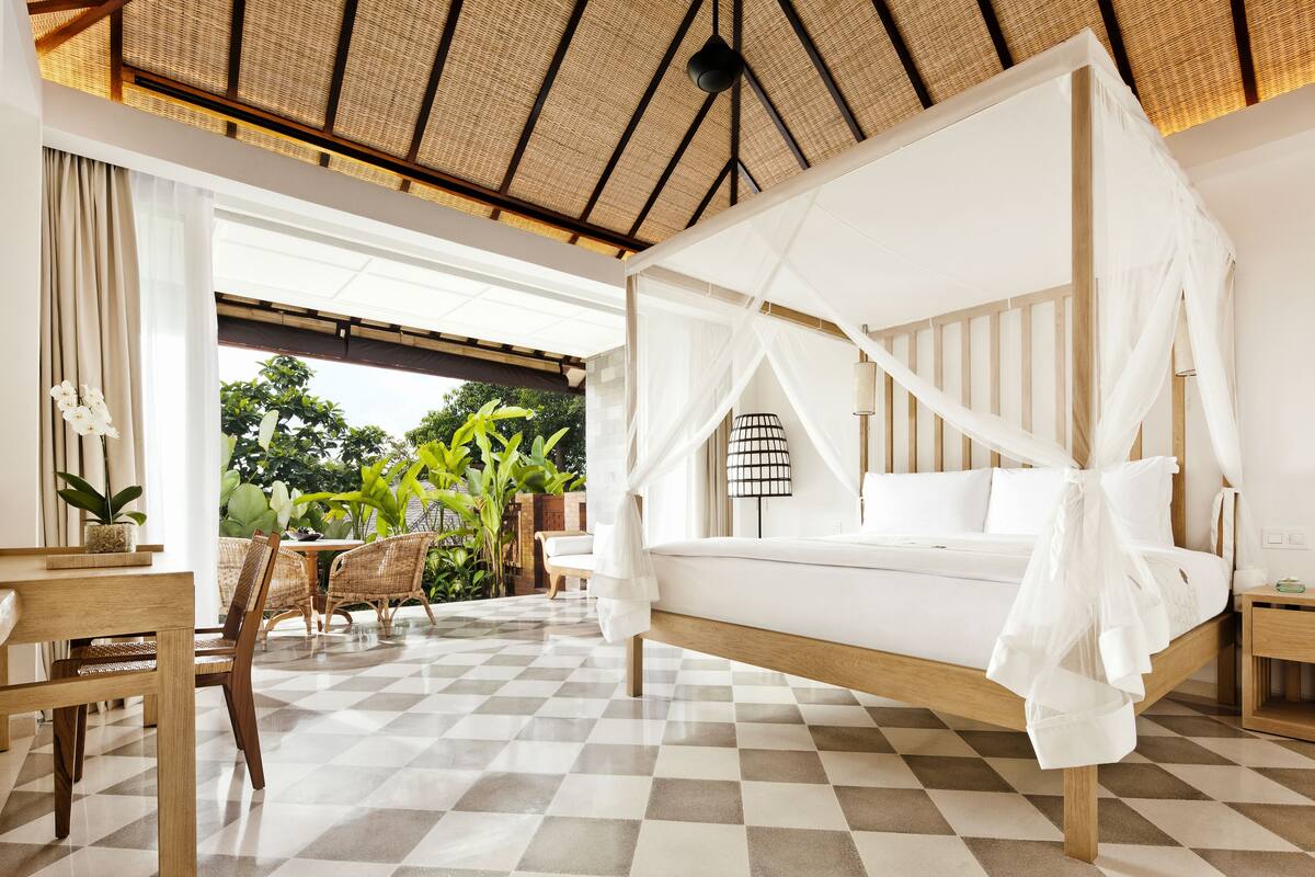 A room with a checkered floor, a bed with a canopy, wicker furniture, and a view of greenery outside.