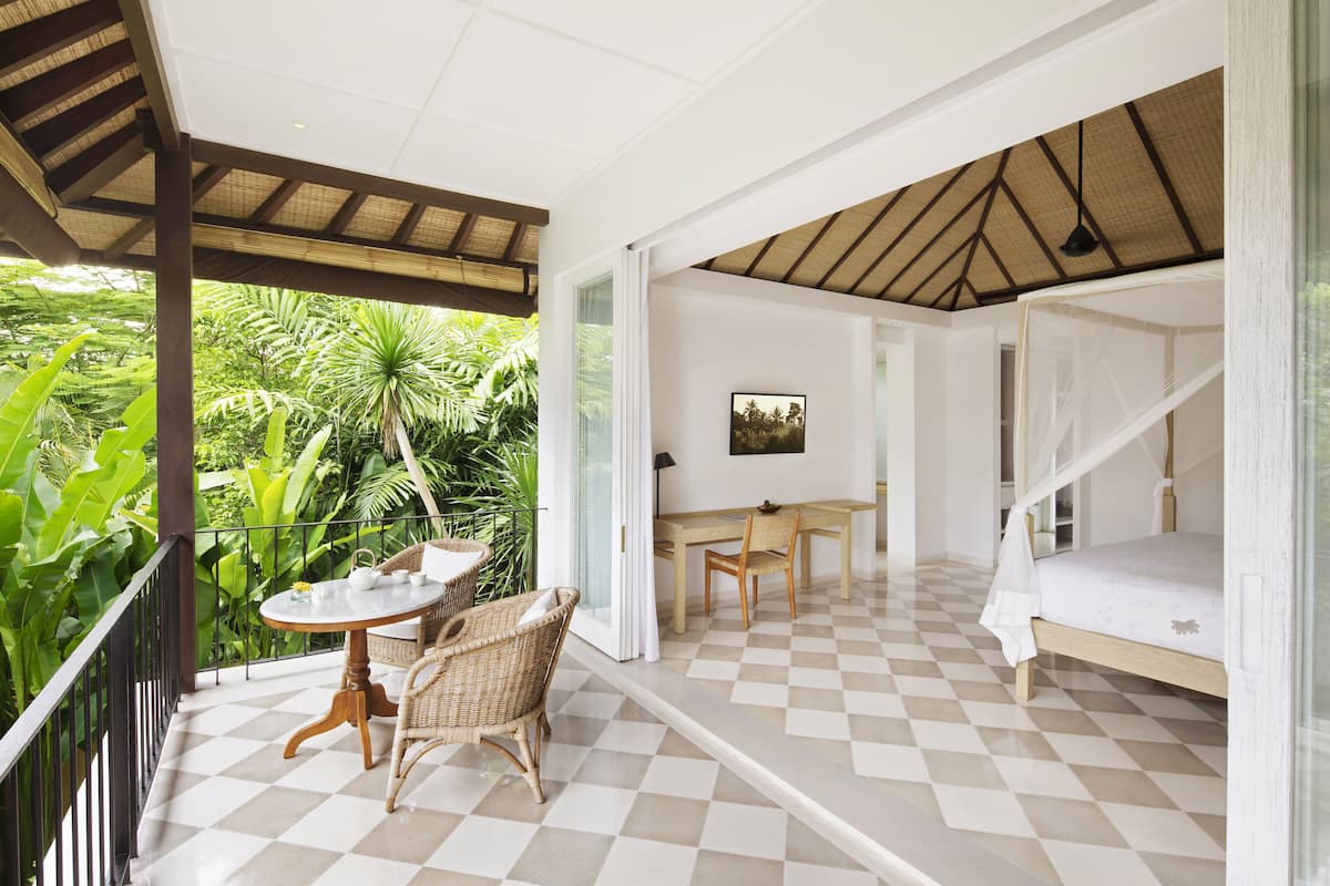 A room with a checkered floor, a bed with a canopy, a dining table with chairs, and a view of lush greenery outside.
