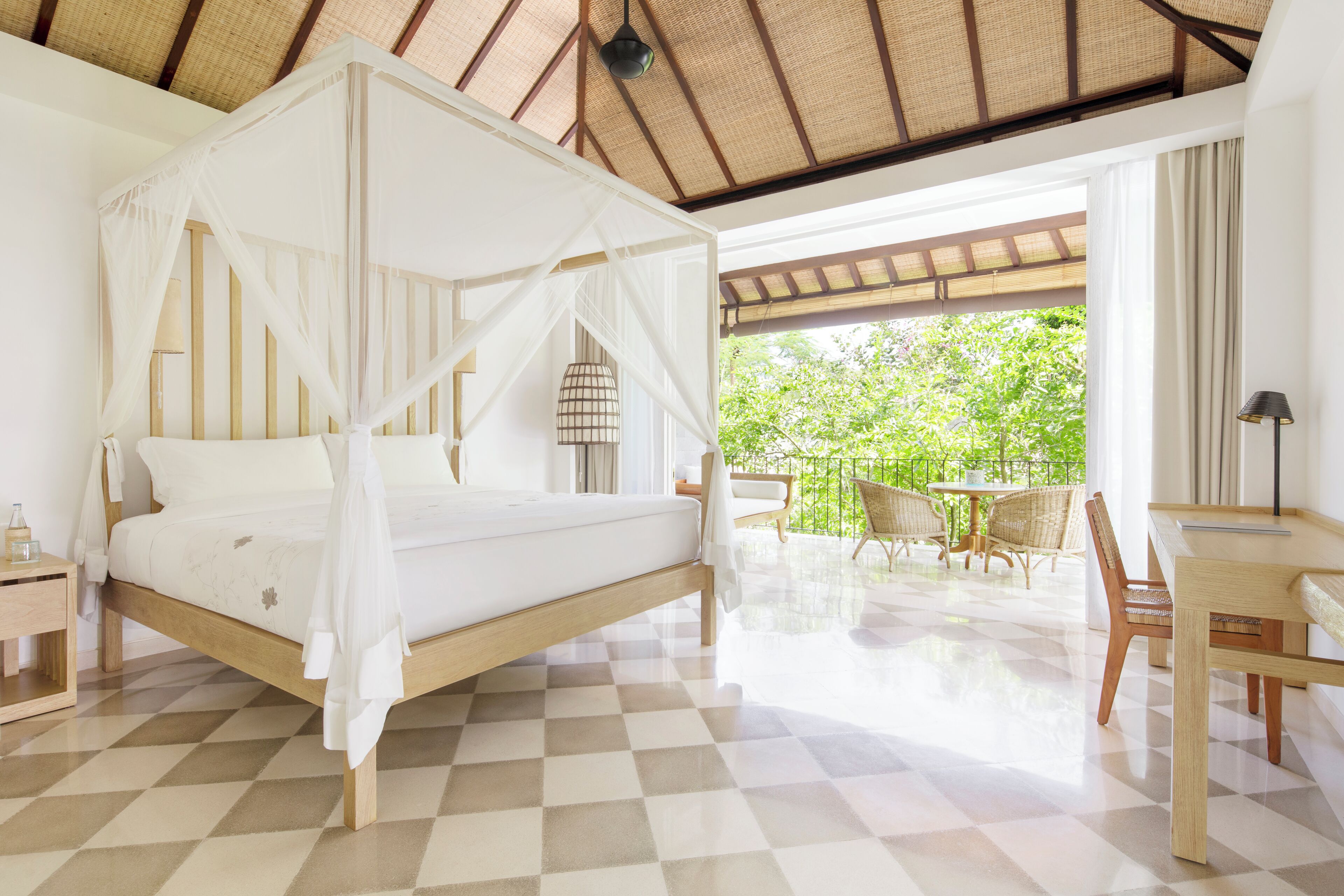 A bedroom with a four-poster bed, a wooden desk, and a view of greenery outside.