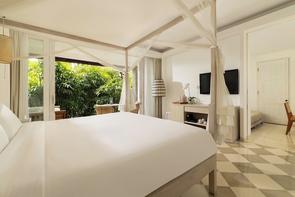 A bedroom with a large bed, a bedside table, a TV, a window with curtains, and a view of greenery outside.