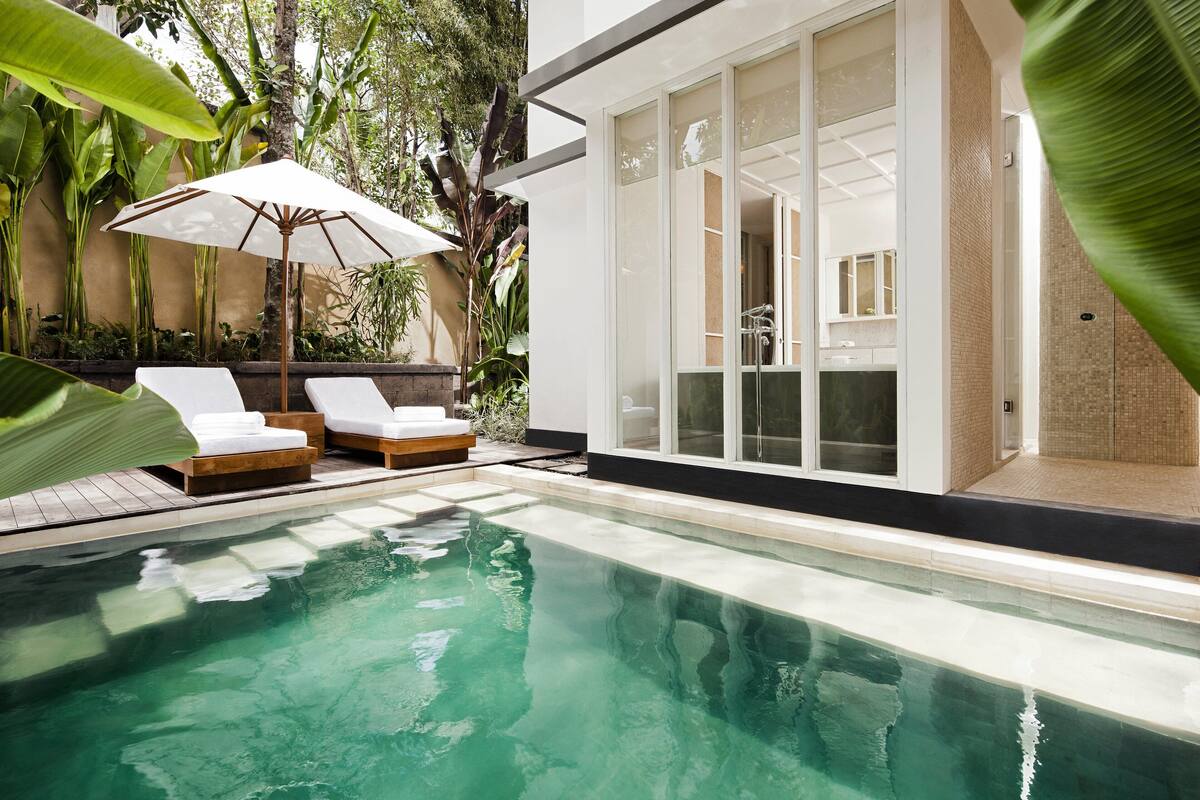 A modern pool area with lounge chairs and a large umbrella, surrounded by lush greenery.