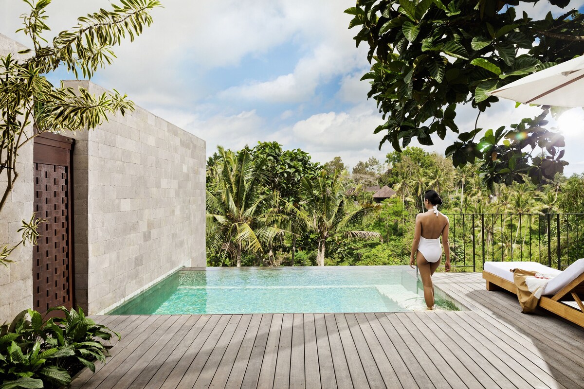 A wooden deck with a swimming pool, a person in a swimsuit, and lush greenery in the background.